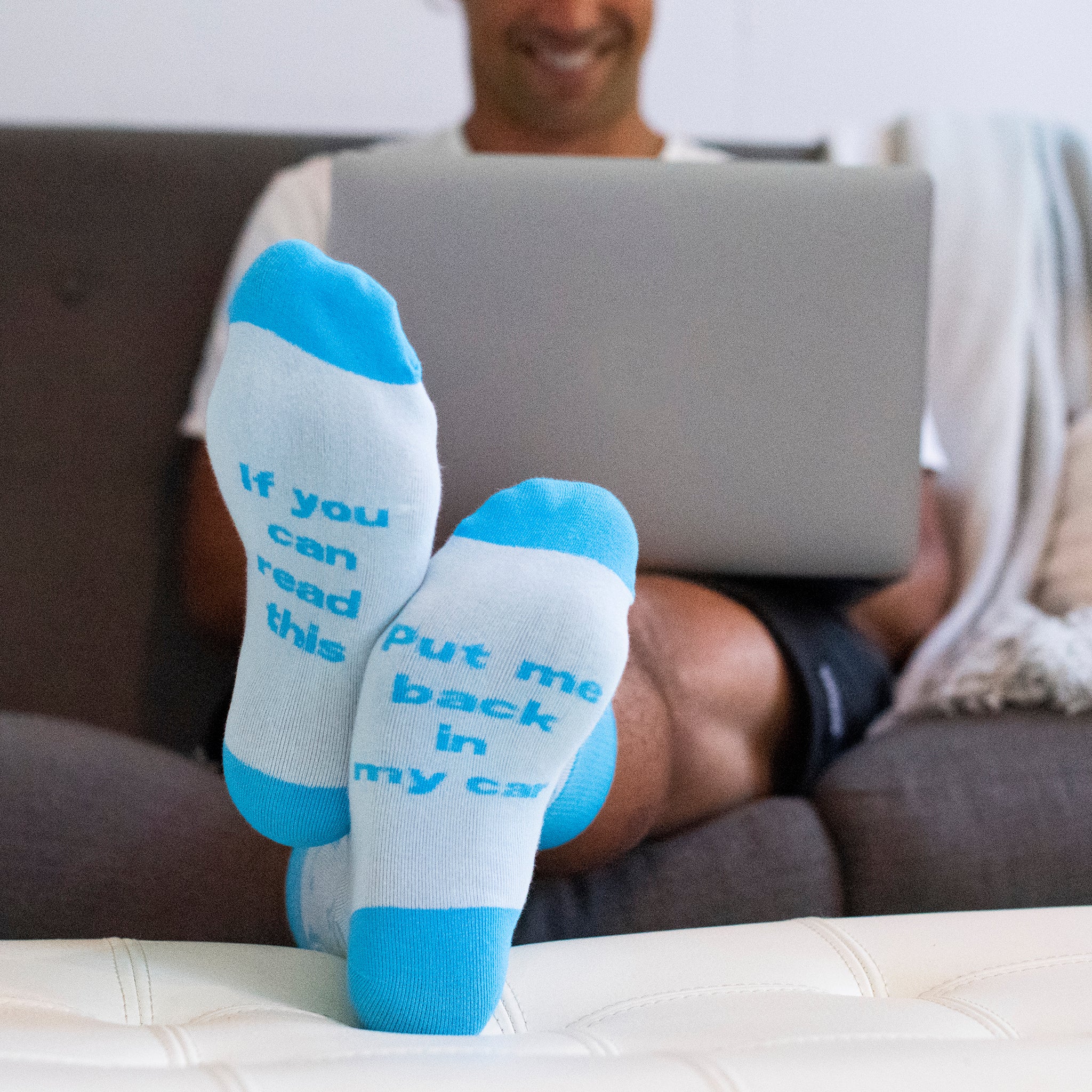 A young man sitting on a couch with a laptop, socked feet resting on an ottoman. The socks read "If you can read this, put me back in my car" in blue. The socks are mainly white with blue tips and heels. Designed exclusively for The Shop by Hagerty.