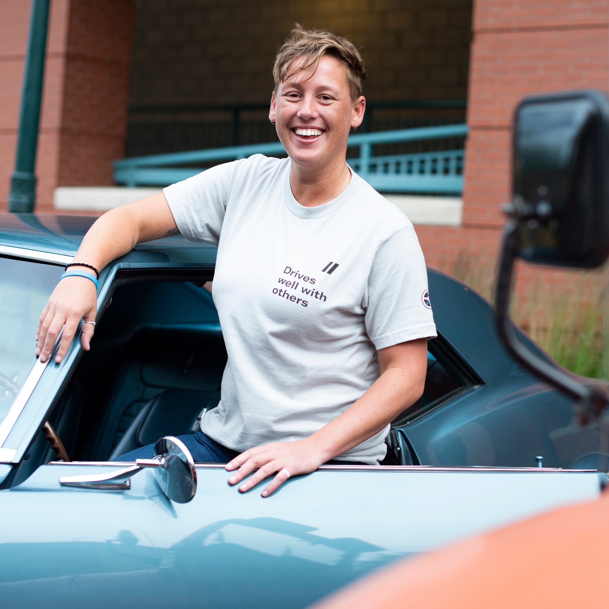A young woman outside the driver's seat of a classic car wearing a light grey Driver's Club T-Shirt with a screen printed racing stripe design and text that reads "Drives well with others." Designed exclusively for The Shop by Hagerty by Anthem Branding.